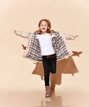 Funny Kid Girl In Opened Beige Raincoat Is Hopping On One Leg With Her Hands Spread Like Wings, Happy Screaming