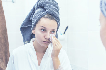 the girl in the bathroom in front of the mirror with a towel on her head applies cream to her face. selective focus. film grain
