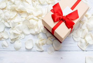 Single gift box out standing from white rose petals on white wooden background. Top view. Copy space on below.