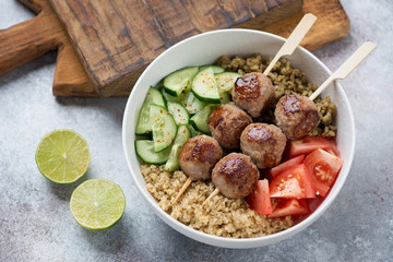 Food bowl with meatball skewers, quinoa, bulgur and fresh vegetables, studio shot on a beige stone background, elevated view