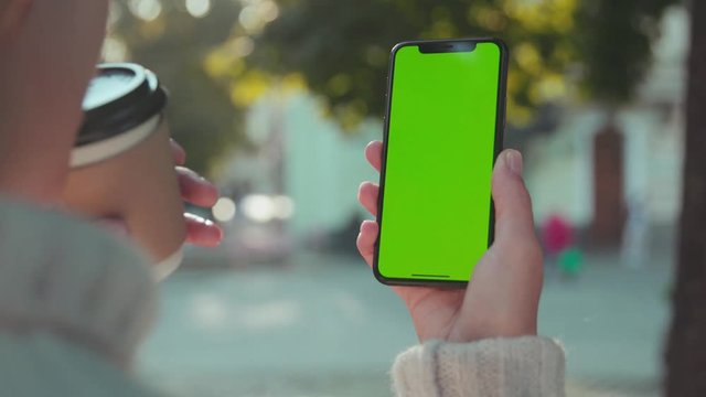 Close-up of young casual woman drinking coffee holding vertical smartphone looking on mock-up phone greenscreen technology in the street.