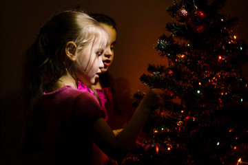 little girls stand near a small Christmas tree in a dark room