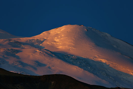 Dawn over the western peak of Mount Elbrus. The snow-covered peaks of a stratovolcano are lit by the rays of the rising sun in the North Caucasus in Russia. © olgapkurguzova
