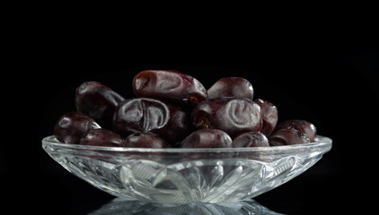 Crystal saucer with a bunch of dried dates on a dark mirror background.