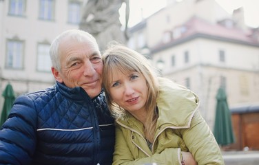 Handsome elderly man is embracing his young blonde wife spending time together outdoors in the ancient city during early spring or autumn.