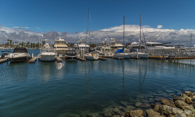 Yachts and boats lined up at the marina with rocks in foreground and clouds in sky in background