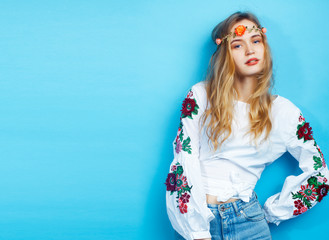 young pretty blond girl posing on blue background, fashion style hippie boho flowers on head