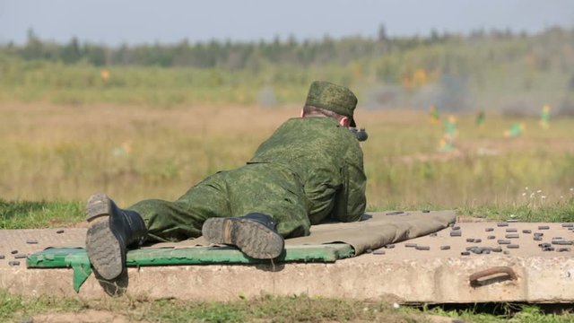 A soldier in a camouflage uniform shoots from a machine gun lying down