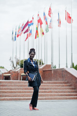 African student female posed with backpack and school items on yard of university, against flags of...