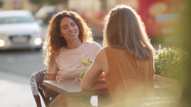 Slowmo medium shot of beautiful young woman with curly hair laughing and chatting with her female friend while sitting at table in outdoor seating area of cafe