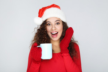 Beautiful american woman wearing santa hat and holding cup of tea on grey background