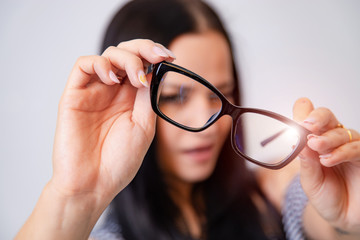 Portrait of a young woman with spectacles in hands. Blurred white background. Girl looks through eyeglasses. Longhaired brunette beautiful girl and black framed eyeglasses. Closeup.