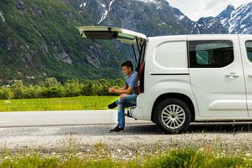 Young travel man sitting in car trunk typing on phone with beauty nature landscape on background