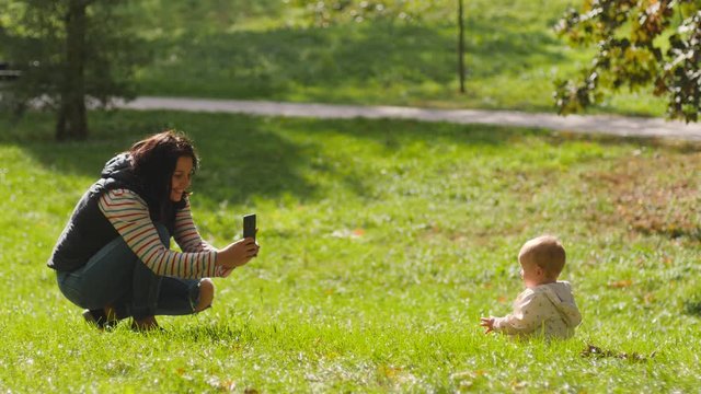 Young mother takes picture of toddler baby child daughter on a park grass via mobile smartphone photo camera for social media