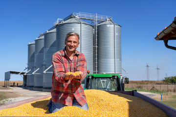 Smiling Farmer Sitting in Tractor Trailer Full of Corn Seeds in front of Farm Grain Bins