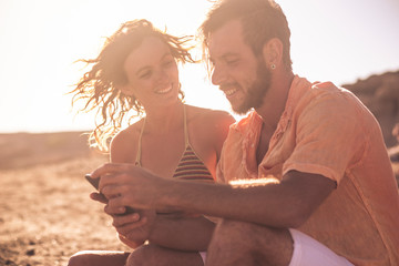 couple of adults at the beach talkin and loking at the phone of the woman sitting on the rocks - woman in bikini looking at ther phone and a man looking at the same phone at the sunset
