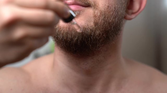 Attractive unshaved man pouring barber oil drops on his beard to soften it. Man doing routine morning procedures at home in front of mirror. Handsome man taking care of his stylish beard. Skincare and