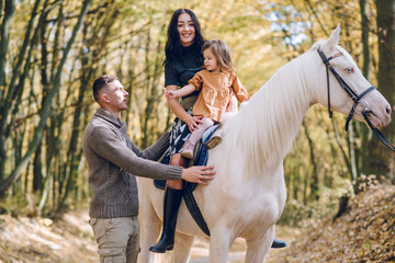 Young family is learning to ride a beautiful white horse in the autumn forest. Riding lesson
