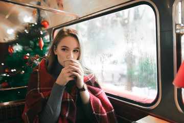 Fototapeta na wymiar Cute girl in plaid on the background of the Christmas tree sitting in a cozy cafe and warmed by a warm drink. Cute lady drinking coffee in a coffee shop and looking away.