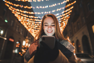 Smiling girl uses smartphone on street background decorated with Christmas lights, looks at smartphone screen and smiles. Street Christmas portrait.