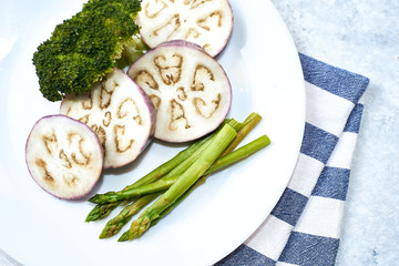 Healthy eating concept. Steamed vegetables in a white plate on a blue table. Eggplant, broccoli, asparagus.