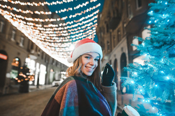 Pretty girl in santa hat smiling smiling and looking at camera. Christmas evening portrait on a decorated street with garlands. New Year concept.