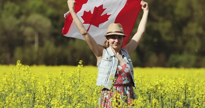 An adult woman with a Canadian flag dances in a rapeseed field in summer.