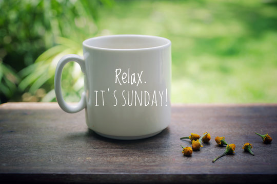 Sunday mug of coffee concept. Morning white coffee with text on it - Relax. It is Sunday, and yellow little flowers arrangement on natural wooden table. Green garden bokeh background.
