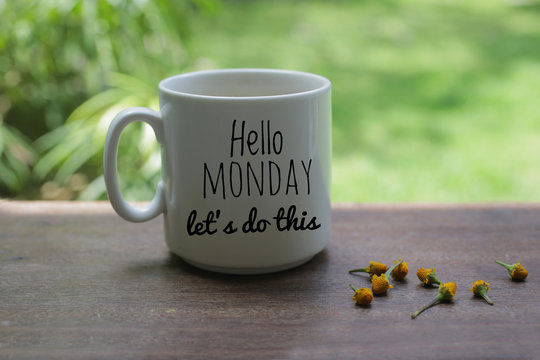 Monday motivational greeting & notes on white mug of coffee - Hello Monday., lets do this. With little yellow flowers arrangement on wooden table and light green garden bokeh background. 