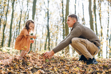Fototapeta premium Family autumn weekend. Young father and his little daughter together in autumn park outdoors. Beautiful baby with leaves on top of dad.