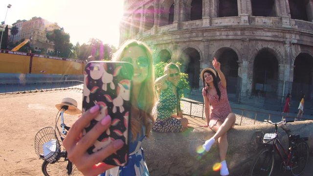 Three happy young women friends tourists with bikes taking videos with smarphone at Colosseum in Rome, Italy at sunrise.