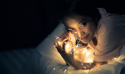 A young woman lying and playing with sparkling lights or fairy light on a white bed In a room with a dim yellow light.