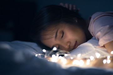 A young woman lying and playing with sparkling lights or fairy light on a white bed In a room with a dim yellow light. She is closing her eyes.