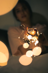 A young woman siting and playing with sparkling lights or fairy light on a white bed In a room with...