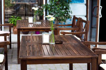 outdoor cafe with wooden tables with a vase of flowers and napkins of brown boards and chairs against the background of the building’s facade and window.