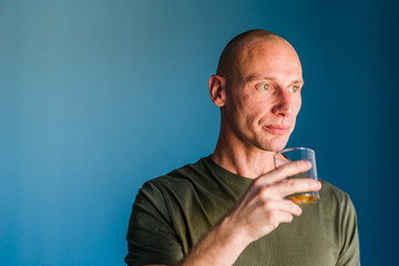 Portrait of young handsome man with short hair holding a glass of whiskey or brandy alcohol drink standing in front of blue wall looking trough the window