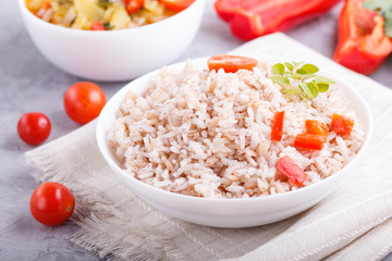 Unpolished rice porridge with stewed vegetables and oregano in white bowl on a gray concrete background. Side view, close up.