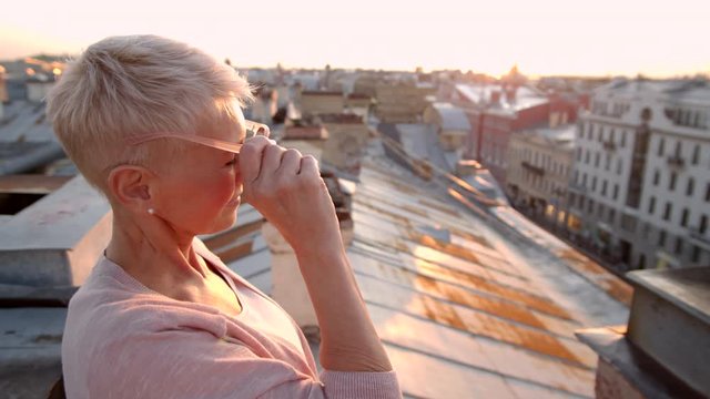 Medium shot of middle-aged woman with short blonde hair standing on roof in downtown, putting off sunglasses and looking at camera