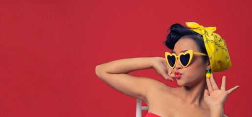 Beautiful young woman wearing vintage yellow glasses and scraf holding earing, kissing lips on red background, Isolated.