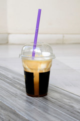Milk brown smoothie in a plastic cup with a lid. The hemisphere-shaped lid on the glass, a purple tube. Glass with coffee on a beige wall background. Delicious cold drinks in disposable dishes.