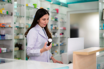 Confident Asian young female pharmacist with smile holding a medicine bottle and searching that product in the computer database in the pharmacy drugstore. Medicine, pharmaceutics, health care