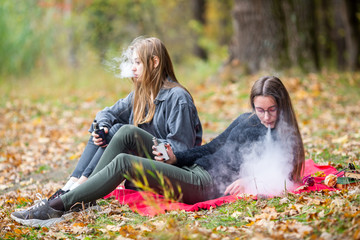 Vape lgbt teenagers. Bisexual lesbian young caucasian teenage girls in casual clothes vape electronic cigarette on the street in the park in the autumn evening. Bad habit.