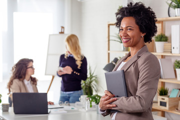 Business woman standing at the office with note book on hand with colegues at the background