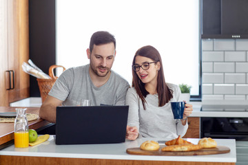 Couple with laptop in pijamas sitting in the kitchen in the morning with pastry and orange juice on table