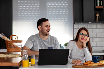 Couple with laptop in pijamas sitting in the kitchen in the morning with pastry and orange juice on table