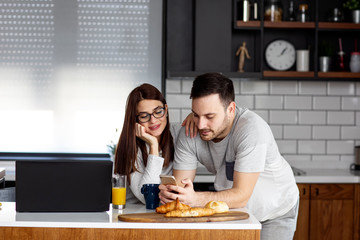 Fototapeta premium Young couple in pijamas eat breakfast at kitchen with laptop beside smile