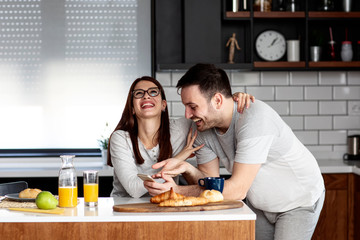 Fototapeta premium Couple in the kitchen eat breakfast with orange juice and pastry at table with mobile phone on hand smile