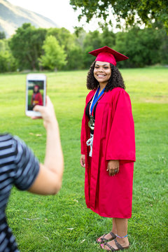 Taking a smartphone photo of a recent high school graduate in her cap and gown. Full length candid photo of a cute smiling diverse girl of African descent.