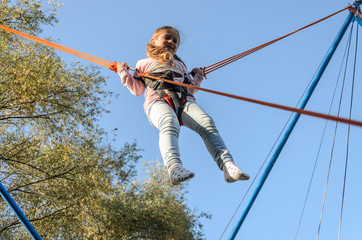 Little charming girl child on elastic ropes jumps on a trampoline in an amusement park
