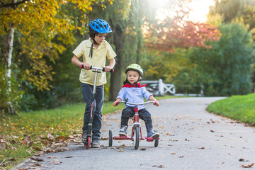 Beautiful blonde two years old toddler boy and his older brother, riding red tricycle and scooter in the park on sunset, beautiful autumn day